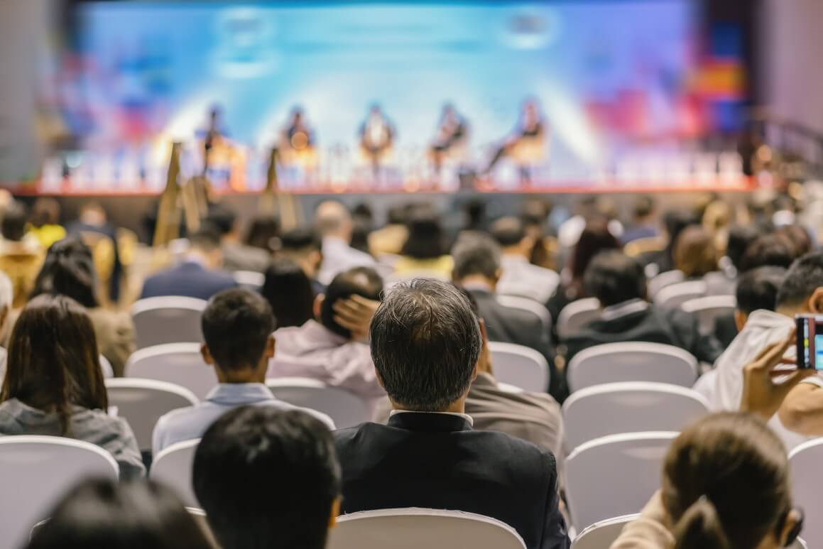 Attendees sitting facing a stage of speakers at a conference.
